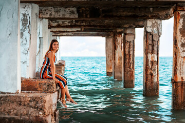 Fototapeta na wymiar Vintage. A smiling woman in a dress sits on the edge of an abandoned old pier, her legs dangling to the sea. The concept of travel and vacation