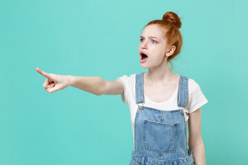 Dissatisfied young readhead girl in casual denim clothes posing isolated on blue turquoise wall background studio. People lifestyle concept. Mock up copy space. Pointing index finger aside, swearing.