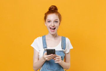 Excited young readhead girl in casual denim clothes white t-shirt isolated on yellow background studio portrait. People lifestyle concept. Mock up copy space. Using mobile phone, typing sms message.