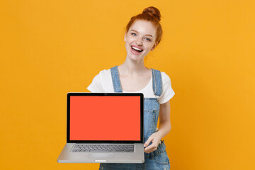 Naklejka premium Funny young readhead girl in casual denim clothes white t-shirt isolated on yellow background in studio. People lifestyle concept. Mock up copy space. Hold laptop pc computer with blank empty screen.