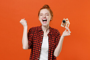 Screaming young readhead girl in casual red checkered shirt posing isolated on orange background studio. People lifestyle concept. Mock up copy space. Hold house bunch of keys doing winner gesture.