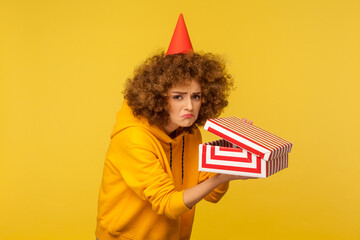 Portrait of unhappy displeased curly-haired woman with funny party cone hat holding opened gift box and looking at camera with disappointment, dissatisfied with present. indoor studio shot, isolated