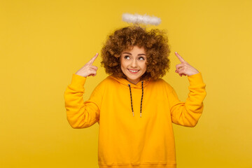 Portrait of charming angel with halo, kind curly-haired hipster woman pointing at saint nimbus over...