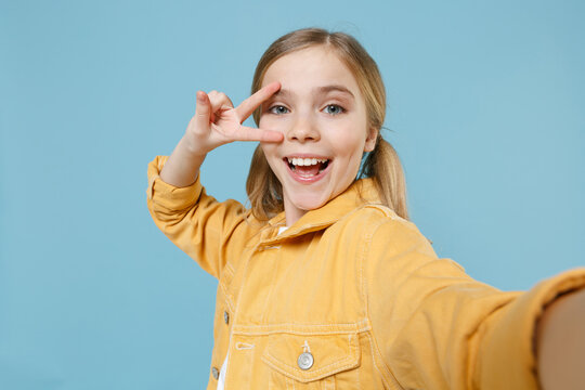 Close up of cheerful little blonde kid girl 12-13 years old in yellow jacket posing isolated on blue background. Childhood lifestyle concept. Doing selfie shot on mobile phone, showing victory sign.
