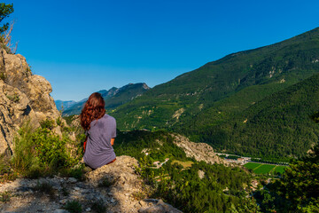 Naklejka premium A full body shot of a thoughtful unrecognizable young Caucasian redhead hiker sitting on a boulder on a hiking path and enjoying the view of the French Alps during sunset