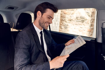 Reading latest news. Handsome young man in full suit reading a newspaper while sitting in the car