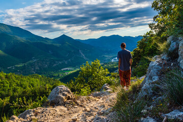 Naklejka premium A full body shot of a thoughtful unrecognizable young Caucasian redhead hiker standing on a hiking path and enjoying the view of the French Alps during sunset