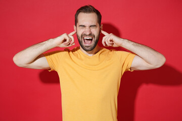 Fototapeta na wymiar Crazy young bearded man guy in casual yellow t-shirt posing isolated on red wall background. People lifestyle concept. Mock up copy space. Covering ears with fingers, keeping eyes closed, screaming.