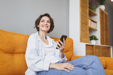 Pretty young woman girl in casual clothes sit on couch spending time in living room at home. Rest relax good mood leisure lifestyle concept. Mock up copy space. Using mobile phone, typing sms message.