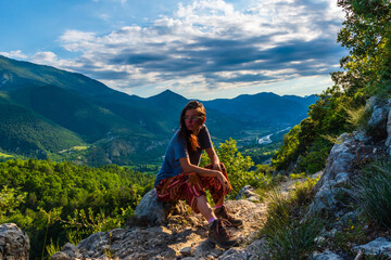 A full body shot of a thoughtful young Caucasian redhead hiker sitting on a boulder on a hiking path and enjoying the view of the French Alps during sunset