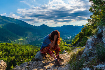 A full body shot of a thoughtful and tired unrecognizable young Caucasian redhead hiker sitting on a boulder on a hiking path and enjoying the view of the French Alps during sunset