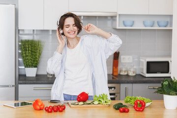 Funny young housewife woman girl in casual clothes preparing vegetable salad cooking food in light kitchen at home. Dieting healthy lifestyle concept. Mock up copy space. Listen music with headphones.