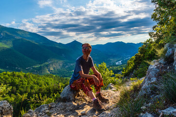 A full body shot of a thoughtful young Caucasian redhead hiker sitting on a boulder on a hiking path and enjoying the view of the French Alps during sunset