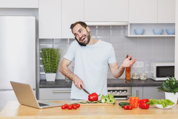 Fototapeta premium Funny young bearded man guy in white casual t-shirt talking on mobile phone preparing vegetable salad cooking food in light kitchen at home. Dieting healthy lifestyle concept. Mock up copy space.