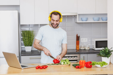 Funny young bearded man guy in white casual t-shirt listen music with headphones preparing vegetable salad cooking food in light kitchen at home. Dieting healthy lifestyle concept. Mock up copy space.