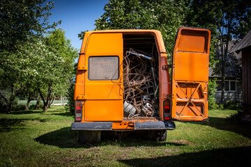 Rear view of an old orange vintage van with the right back door opened filled with scrape metal, waiting to be recycled in a natural green yard with trees and lawn under the summer sun © J S