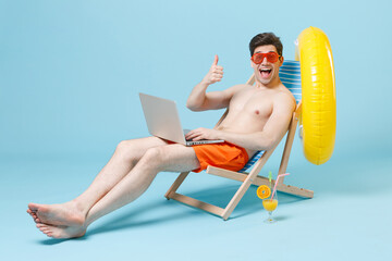 Excited young man guy in orange shorts glasses sit on deck chair isolated on blue background...
