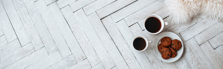 Breakfast in the morning. Two white cups of coffee and chocolate chip cookies stand on a light wooden parquet and pillows lie nearby. Homeliness, day off. Pastel colors.