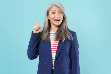 Excited gray-haired business woman in blue suit posing isolated on blue background studio. Achievement career wealth business concept. Mock up copy space. Holding index finger up with great new idea.