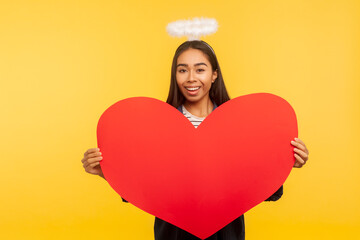 Portrait of beautiful angelic girl with holy nimbus holding big read heart and smiling to camera,...