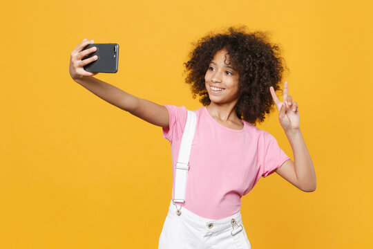 Funny african american kid girl 12-13 years old in pink t-shirt isolated on yellow background. Childhood lifestyle concept. Mock up copy space. Doing selfie shot on mobile phone, showing victory sign.
