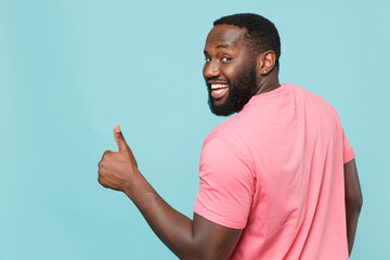 Fototapeta premium Back rear view of funny young african american man guy in casual pink t-shirt posing isolated on blue background studio. People lifestyle concept. Mock up copy space. Showing thumb up, looking camera.
