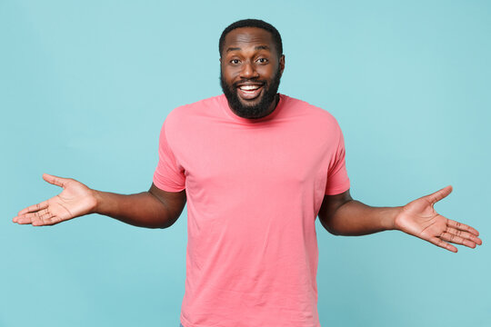 Smiling smirked young african american man guy in casual pink t-shirt isolated on blue wall background studio portrait. People sincere emotions lifestyle concept. Mock up copy space. Spreading hands.
