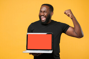 Joyful african american man in casual black t-shirt isolated on yellow background. People lifestyle concept. Mock up copy space. Hold laptop pc computer with blank empty screen doing winner gesture.