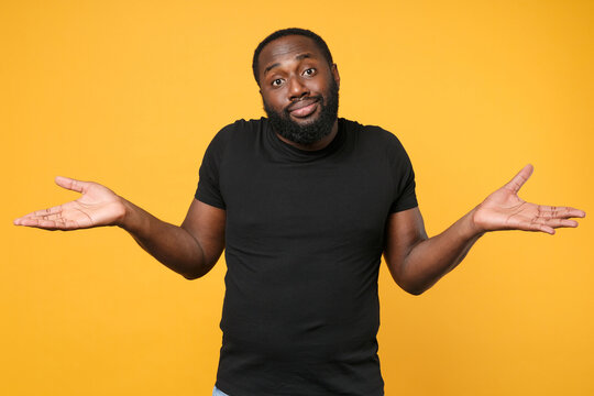 Confused african american man football fan in casual black t-shirt isolated on yellow wall background studio portrait. People sincere emotions lifestyle concept. Mock up copy space. Spreading hands.