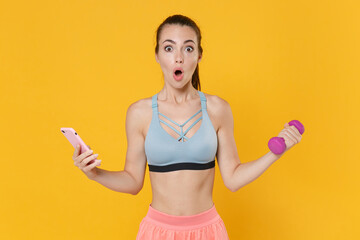 Shocked young fitness sporty woman girl in sportswear working out isolated on yellow background portrait. Workout sport motivation lifestyle concept. Doing exercise with dumbbells, using mobile phone.