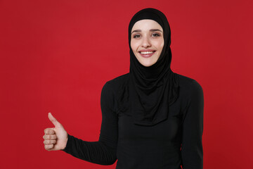 Smiling young arabian muslim woman in hijab black clothes posing isolated on bright red wall background, studio portrait. People religious lifestyle concept. Mock up copy space. Showing thumb up.