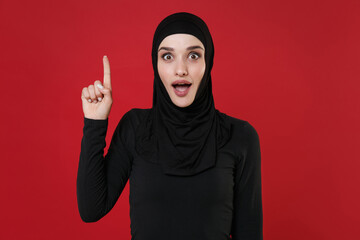 Excited young arabian muslim woman in hijab black clothes posing isolated on red background studio. People religious lifestyle concept. Mock up copy space. Holding index finger up with great new idea.