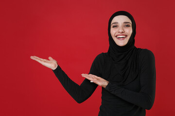 Cheerful young arabian muslim woman in hijab black clothes posing isolated on bright red wall background studio portrait. People religious lifestyle concept. Mock up copy space. Pointing hands aside.