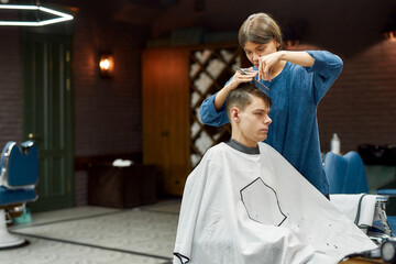 Serving client in barbershop. Professional female hair stylist making trendy haircut for a young caucasian guy, she is using metal shears and comb