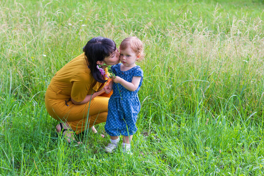 a young mother kisses her little daughter on the cheek with a pink flower in her hand in a meadow with green grass in a Park on a summer day