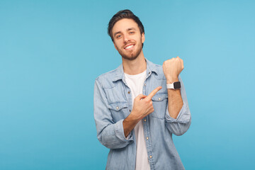 Please, hurry up! Portrait of cheerful punctual man in worker denim shirt pointing at wrist watch and smiling, showing smartwatch devise with mock up display. indoor isolated on blue background