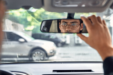Close up back view portrait of man in specs adjusting rearview mirror while sitting in his car, selective focus