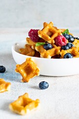 Tiny (Mini) waffles for breakfast with berries in a white bowl on a light background.