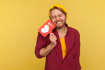 Extremely excited hipster guy in checkered shirt holding Heart Like counter button, smiling with pleasure expression, rejoicing popularity on Internet, social network. studio shot isolated on yellow