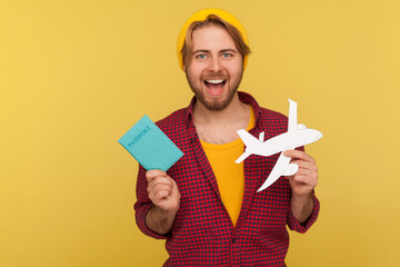 Positive hipster tourist guy in beanie hat, checkered shirt holding paper plane and passport,...