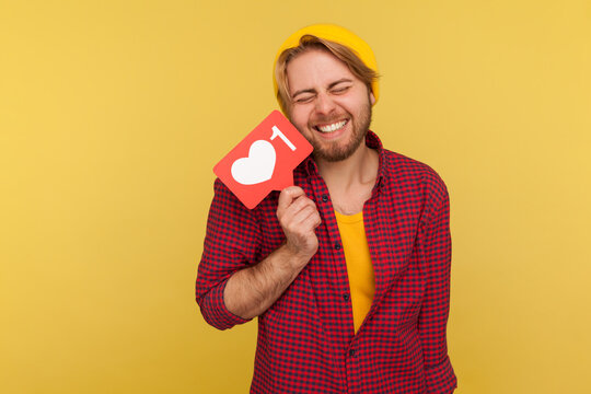 Extremely excited hipster guy in checkered shirt holding Heart Like counter button, smiling with pleasure expression, rejoicing popularity on Internet, social network. studio shot isolated on yellow