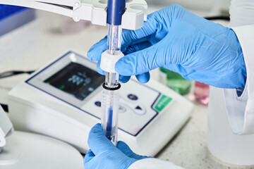 Woman in a rubber gloves holding a glass electrode for measuring of pH of the solution using pH meter. Analytical or electro chemistry laboratory. 