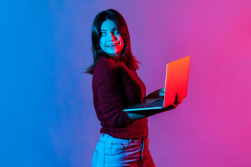 Neon light portrait of successful joyful office worker girl looking away with happy pensive face, smiling holding laptop, typing on keyboard, doing freelance job. colorful neon light, studio shot - obrazy, fototapety, plakaty