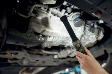 Vehicle problems. Close up shot of hand of mechanic checking car in auto service with lifted vehicle using flashlight. Car service, repair, maintenance concept