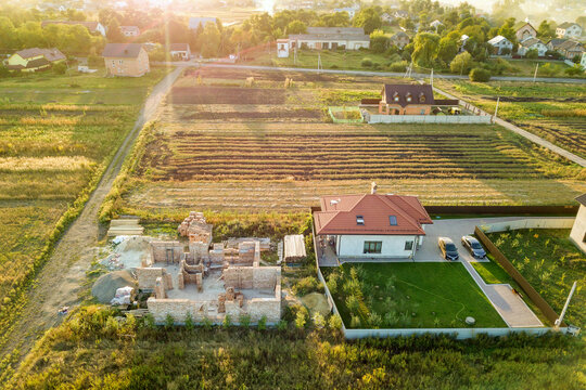 Top down aerial view of two private houses, one under construction with concrete foundament and brick walls and another finished with red tiled roof.