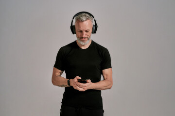 Middle aged muscular man in black t shirt and headphones using smartphone while posing in studio over grey background