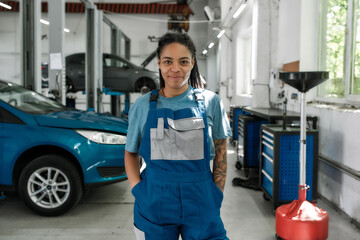 Come for help. Portrait of young african american woman, professional female mechanic in uniform smiling at camera, standing in auto repair shop. Car service, repair, maintenance and people concept
