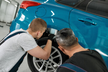 Quality with new standards. Two male mechanics using torch for inspecting wheel of lifted car at auto repair shop