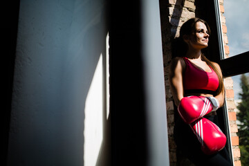 Sport girl in red top standing against the wall and looking out the window. Woman athlete with Boxing gloves.