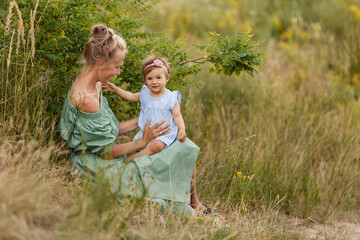 Mother and little daughter walk in blooming summer meadow. Mom loves her child. Toddler girl in Mom's hands. Happy family in beautiful summer day in flowering grass. Tender relations. Cotton outfit